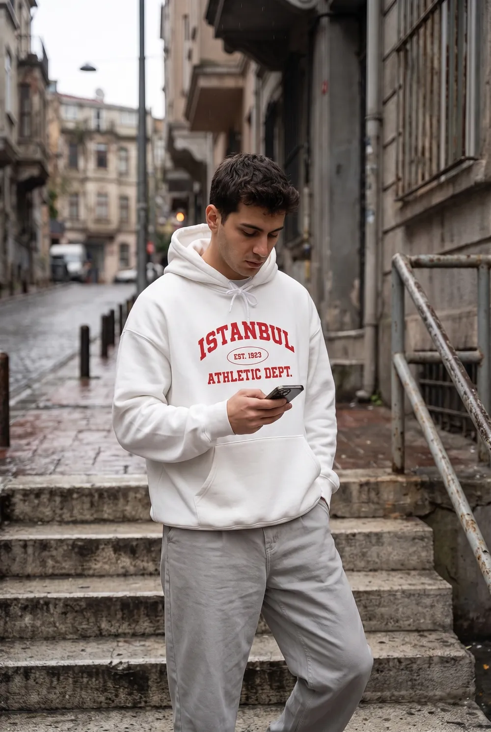 ISTANBUL Athletic Dept. yazılı kırmızı kolej baskılı beyaz hoodie giyen genç adam, taş merdivende