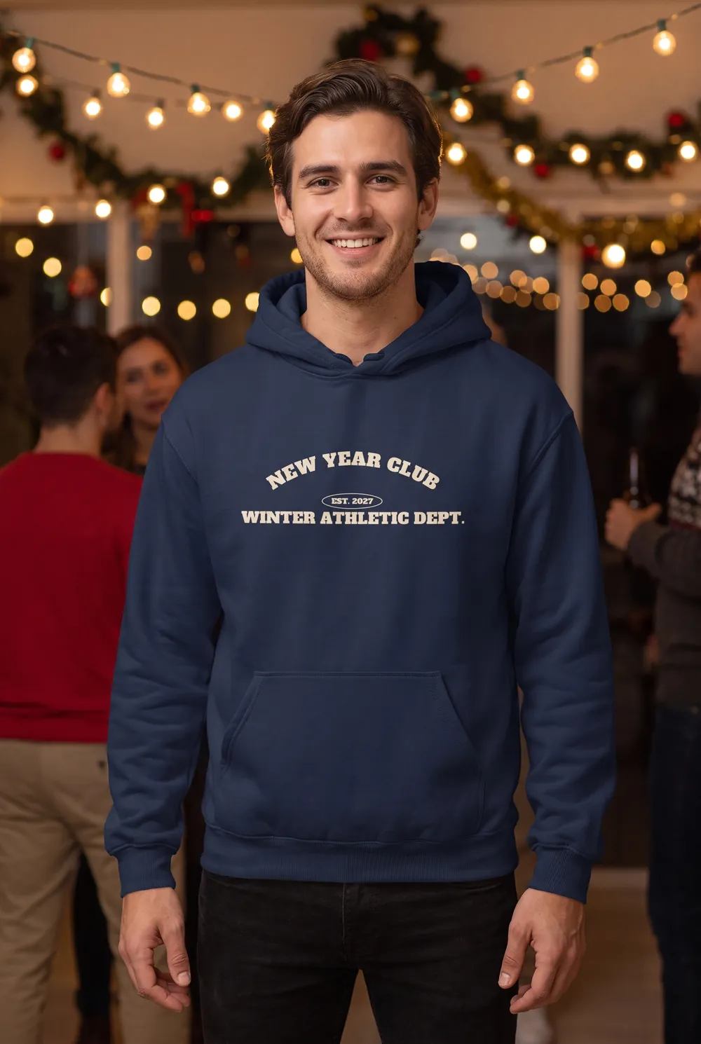 NEW YEAR CLUB Winter Athletic Dept. yazılı lacivert hoodie giyen genç adam, arkada yılbaşı ışıkları
